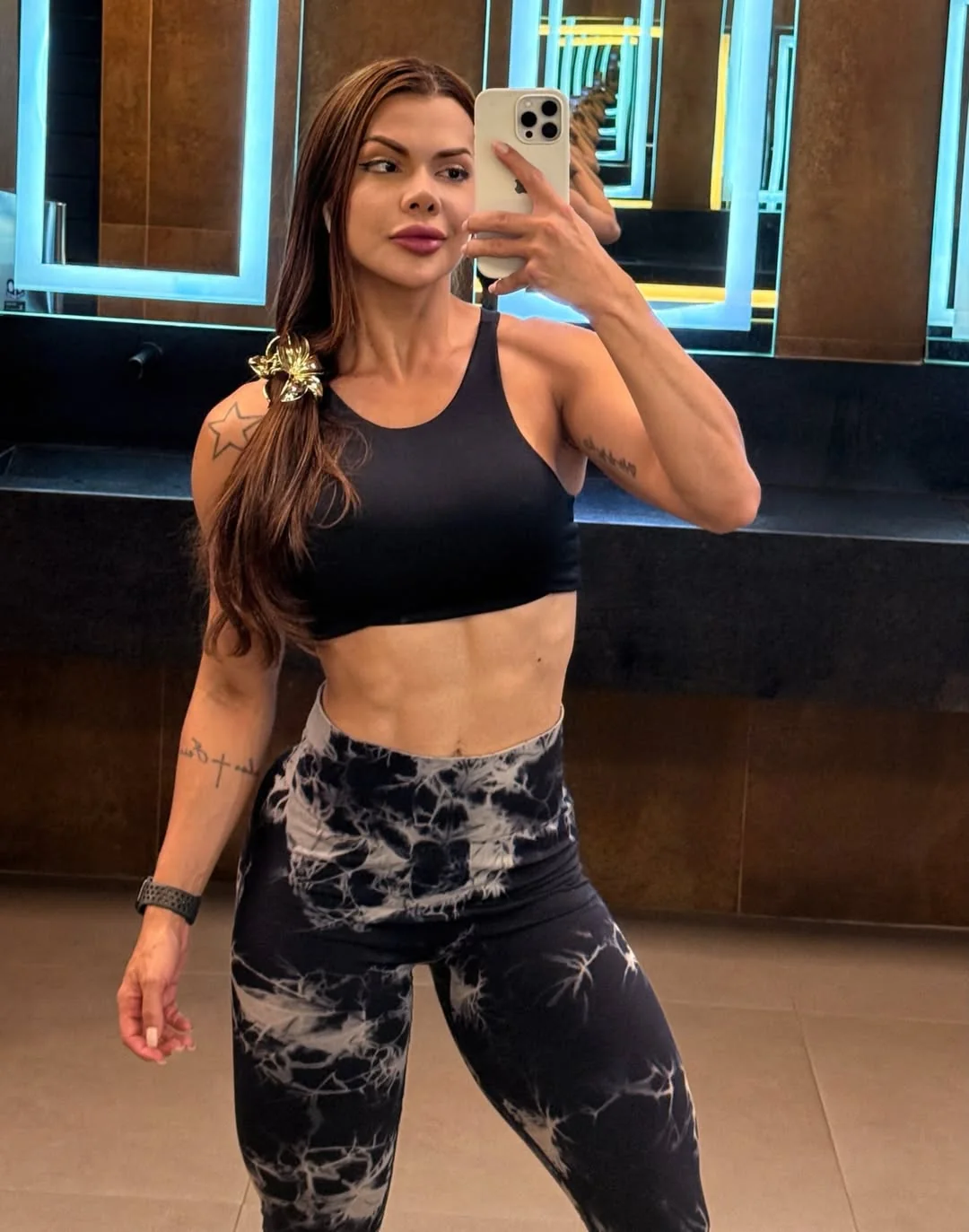 Viviane Andrade em selfie de espelho na academia, de top preto e legging preta com estampa tie-dye, com o cabelo preso de lado
